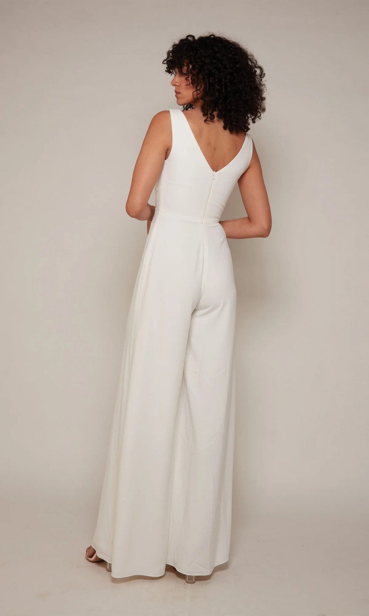 A woman with curly hair stands facing away, wearing the Long White Jumpsuit: Alyce 70052—a sleeveless wide-leg bridal jumpsuit with a chic V-shaped back.